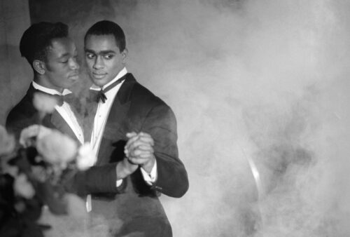 Two Black men, in tuxedos, clasp hands and dance in a smoky foreground in a scene from "Looking for Langston," the 1989 film that reevaluated gay and lesbian contributions to the Harlem Renaissance.