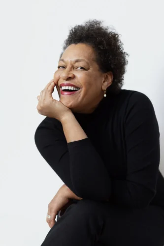 Carrie Mae Weems smiles and poses with left hand resting on her chin. 