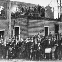Black and white photo of white men with guns in front of a burned building that once was the Record newspaper building